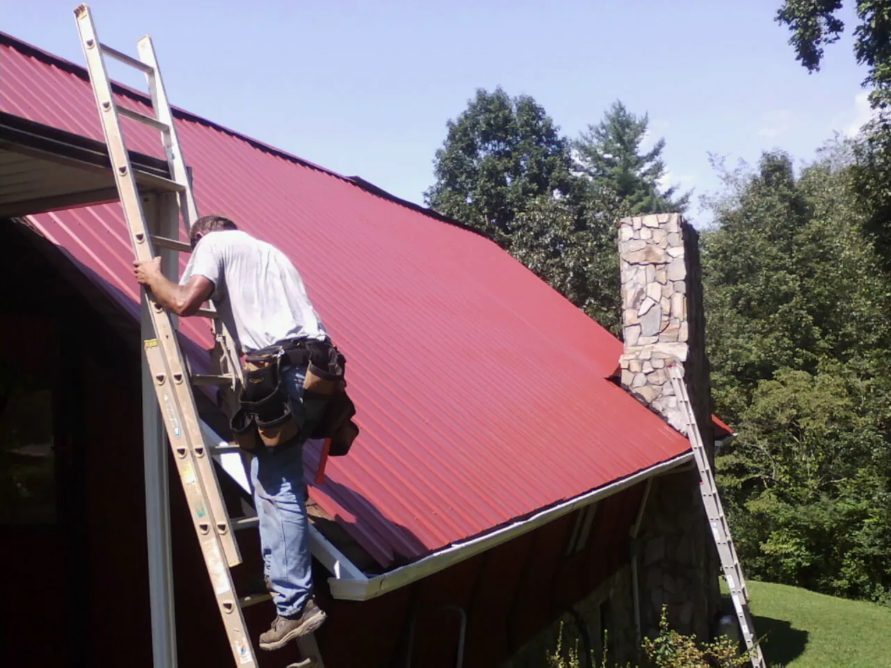 Roofer on a ladder beside a red metal roof and stone chimney — Soffit & Fascia Repair by Roofing Brothers in Dumfries